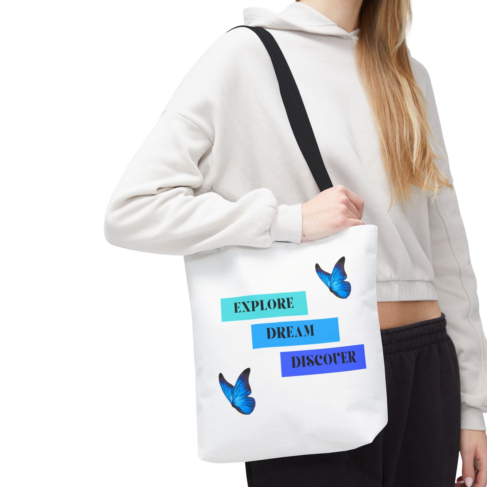 Woman carrying Charming white Explore, Dream, Discover tote bag with black handles, featuring a positive, butterfly-themed design. Made from 100% polyester, this tote is perfect for adventurers and creatives. Available in sizes 13"x13", 16"x16", 18"x18". Luminous Gem Designs.