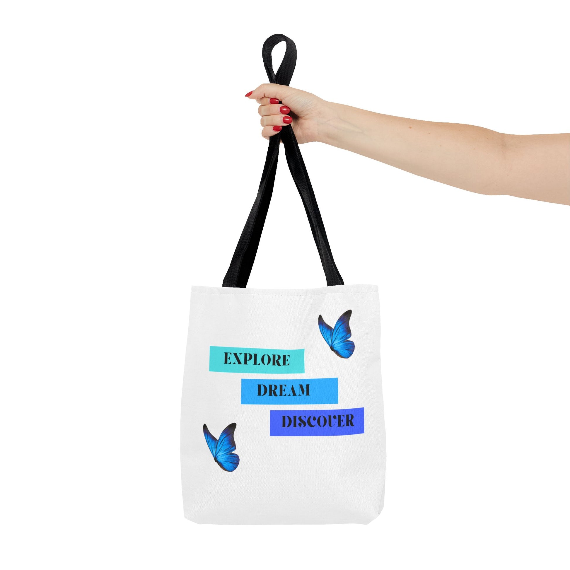 Hand holding Charming white Explore, Dream, Discover tote bag with black handles, featuring a positive, butterfly-themed design. Made from 100% polyester, this tote is perfect for adventurers and creatives. Available in sizes 13"x13", 16"x16", 18"x18". Luminous Gem Designs.